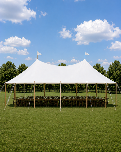 A large white event tent with a peaked roof is set up on a green lawn under a blue sky with white clouds. Long tables and chairs are arranged underneath, with trees lining the background behind the tent.