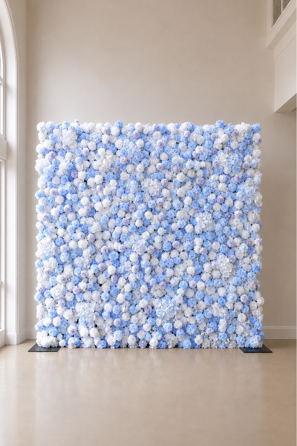 A large decorative wall panel made of dense clusters of light blue and white pom-pom flowers, standing on a polished beige floor in a minimalist indoor room with cream walls and a window on the left.