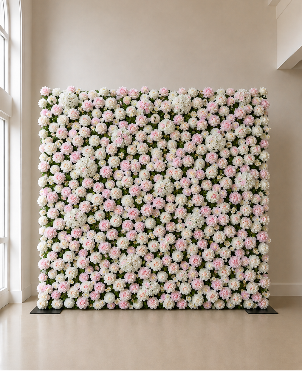 A large rectangular floral wall backdrop covered in dense clusters of white and light pink flowers with green foliage, displayed indoors against a beige wall and floor.