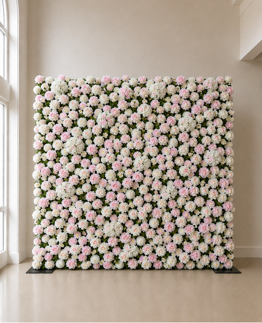 A large rectangular floral wall backdrop covered in dense clusters of white and light pink flowers with green foliage, displayed indoors against a beige wall and floor.